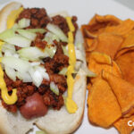 Rudys Hot Dog is a coney style hot dog topped with onions and mustard. photo shows hot dog with sweet potato chips.