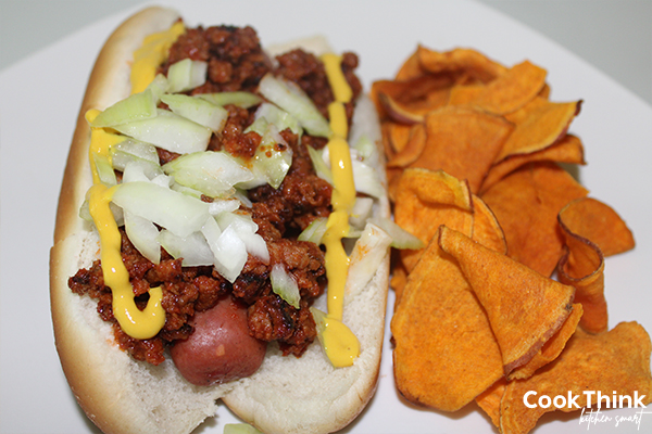 Rudys Hot Dog is a coney style hot dog topped with onions and mustard. photo shows hot dog with sweet potato chips.